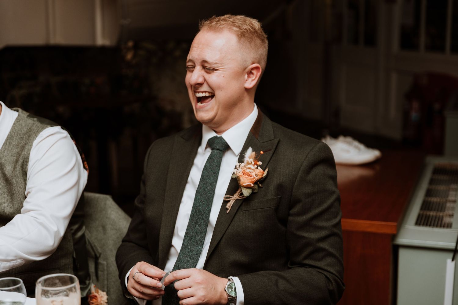 Wedding guest in dark suit with orange boutonniere laughs during reception celebration.