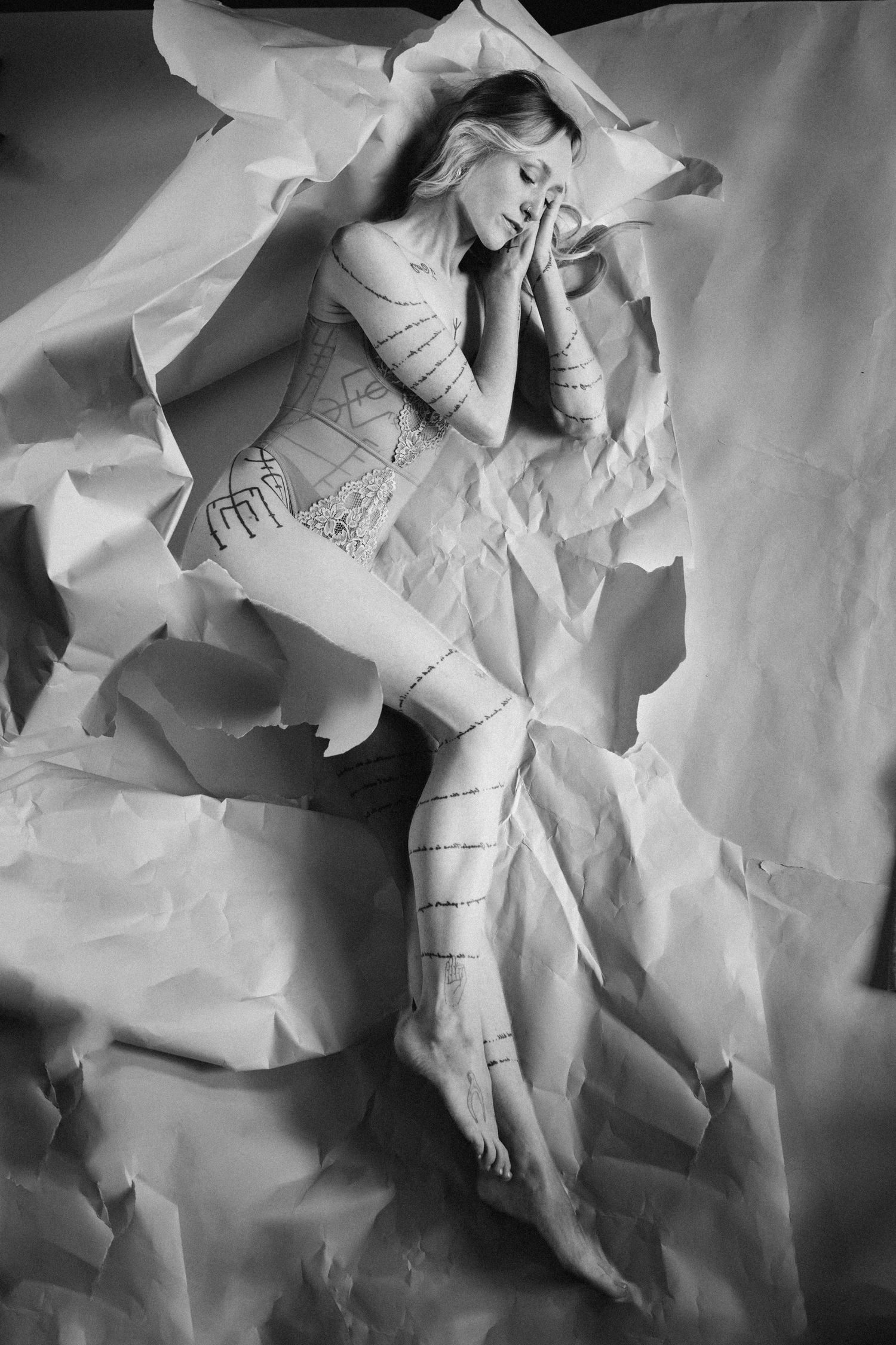 Black and white overhead shot of a woman in a painted bodysuit lying on crumpled paper.
