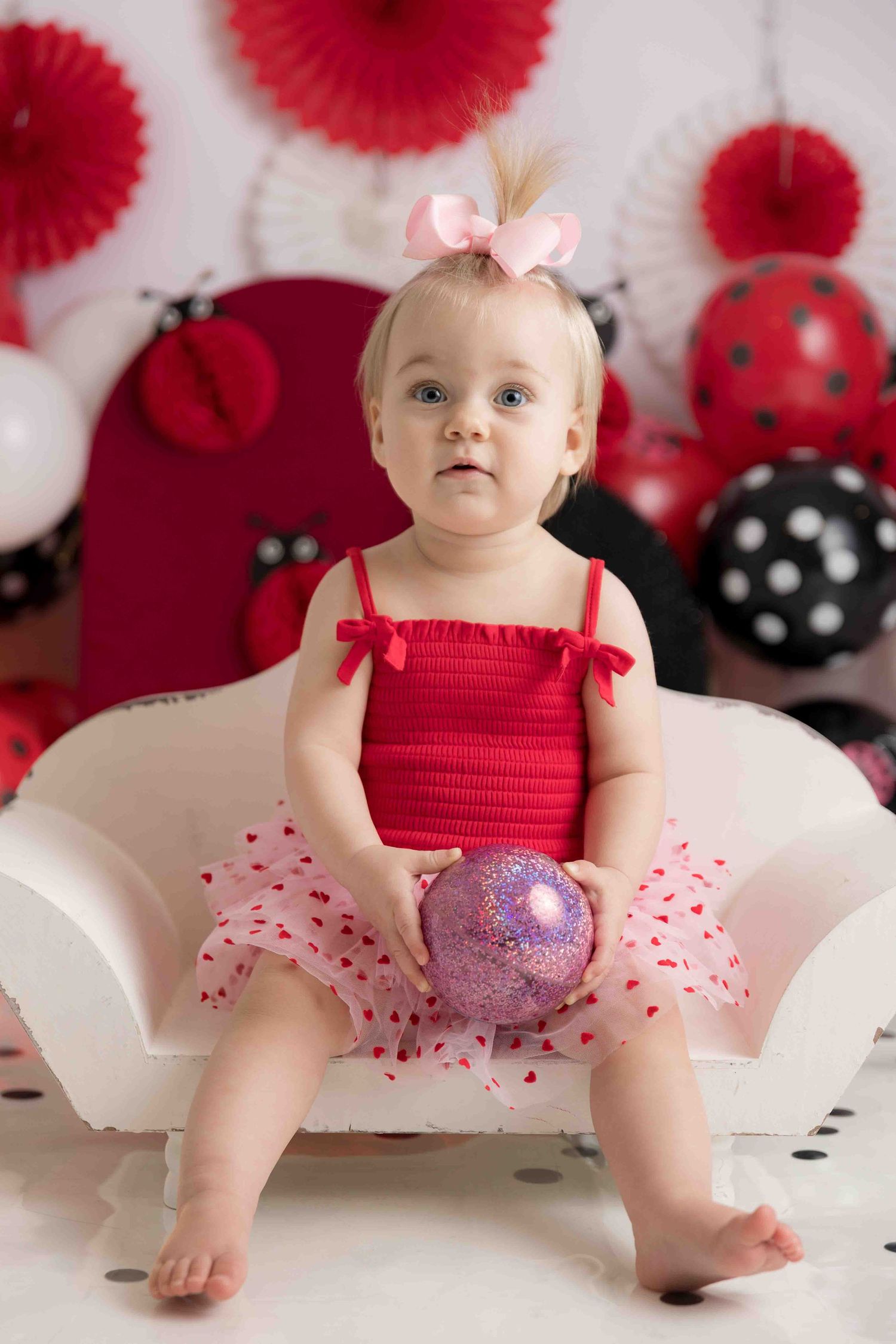 A baby in a red and pink tulle outfit sits against a backdrop with red paper decorations and polka dot accents.