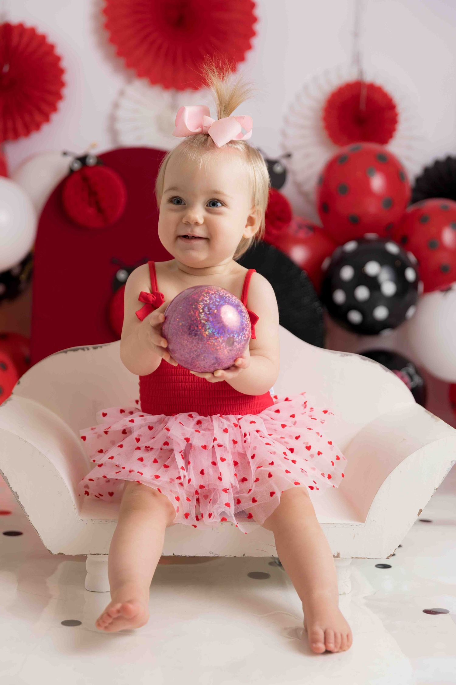 A baby in a pink tutu dress poses against a white backdrop with red paper fans and polka dot decorations.