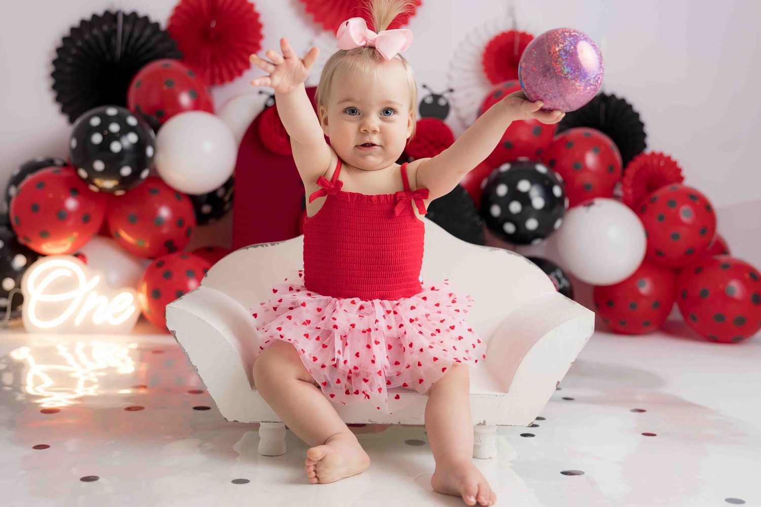A small child in a red and white tutu dress sits in front of a festive balloon arch with red and black decorations.