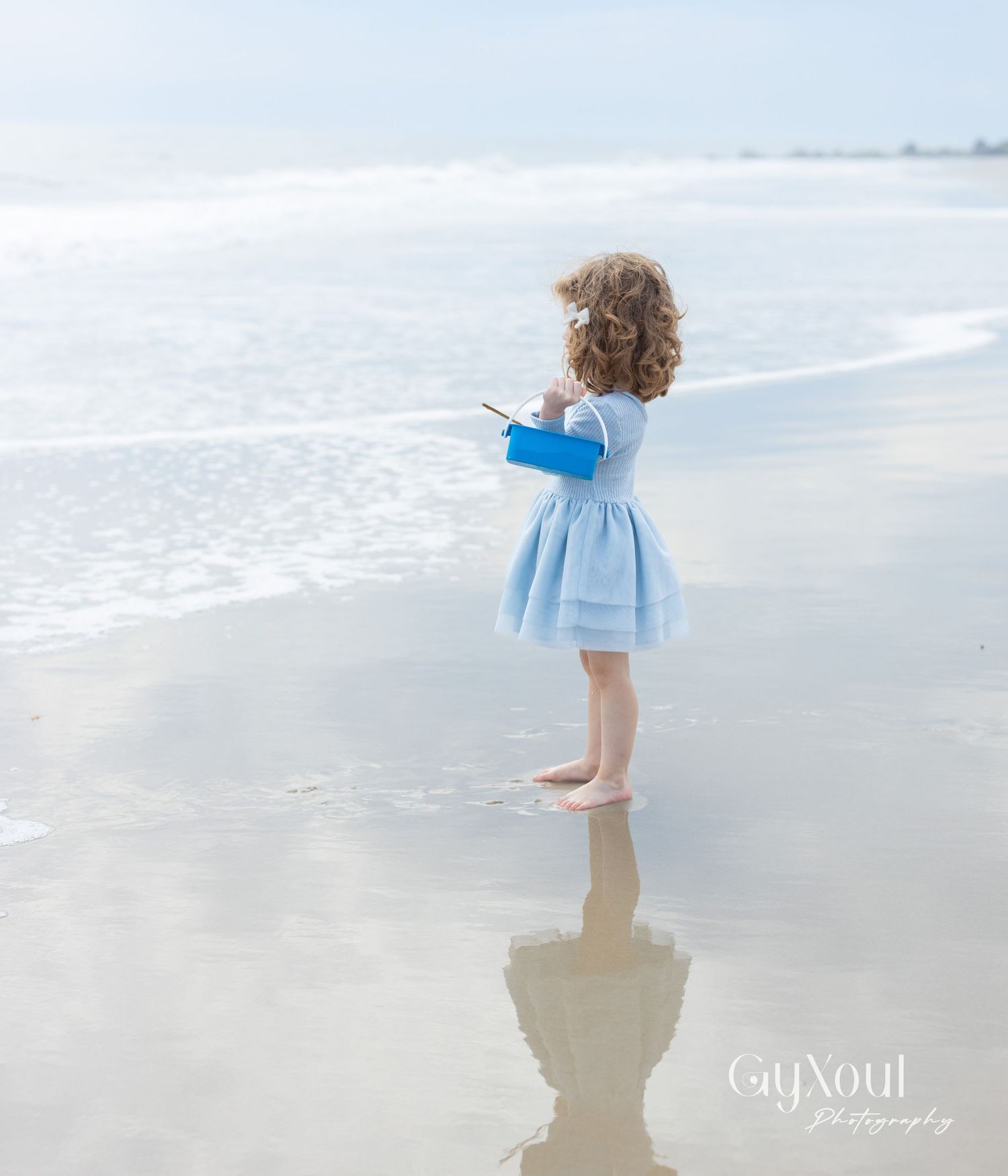 A small child in a blue dress stands at the edge of the ocean surf, their reflection visible in the wet sand.