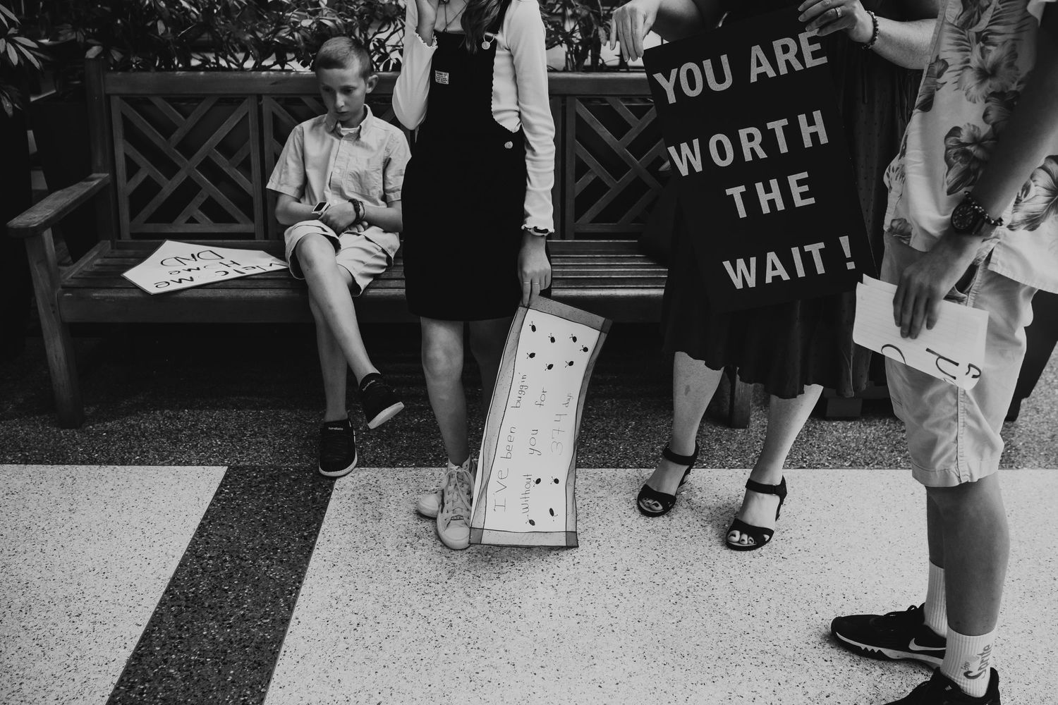 Black and white photo of people waiting in line by a sign that reads 'YOU ARE WORTH THE WAIT' in classic vintage style.