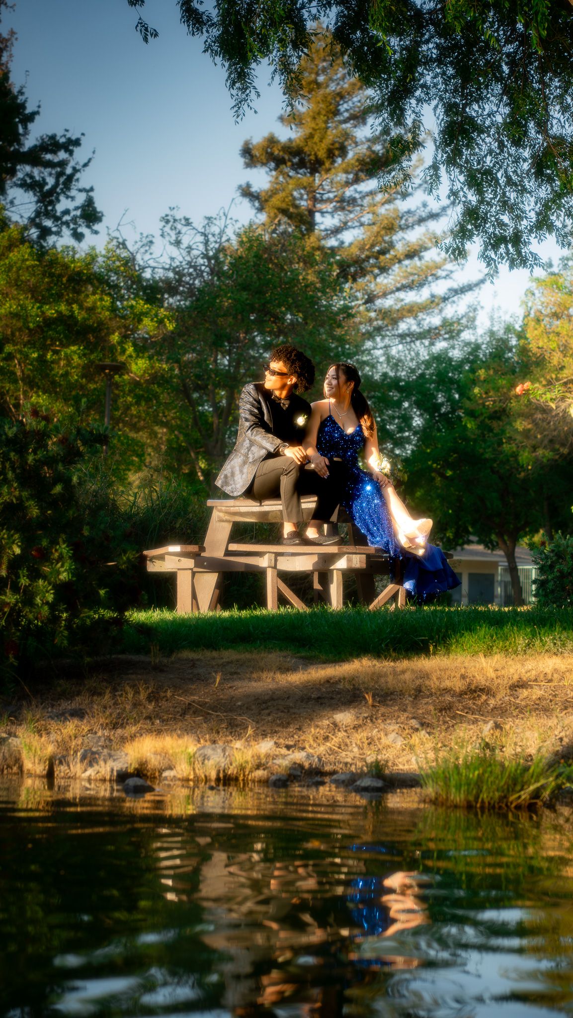 A couple in formal attire sits on a wooden platform by a tranquil pond with their reflection visible in the water below.