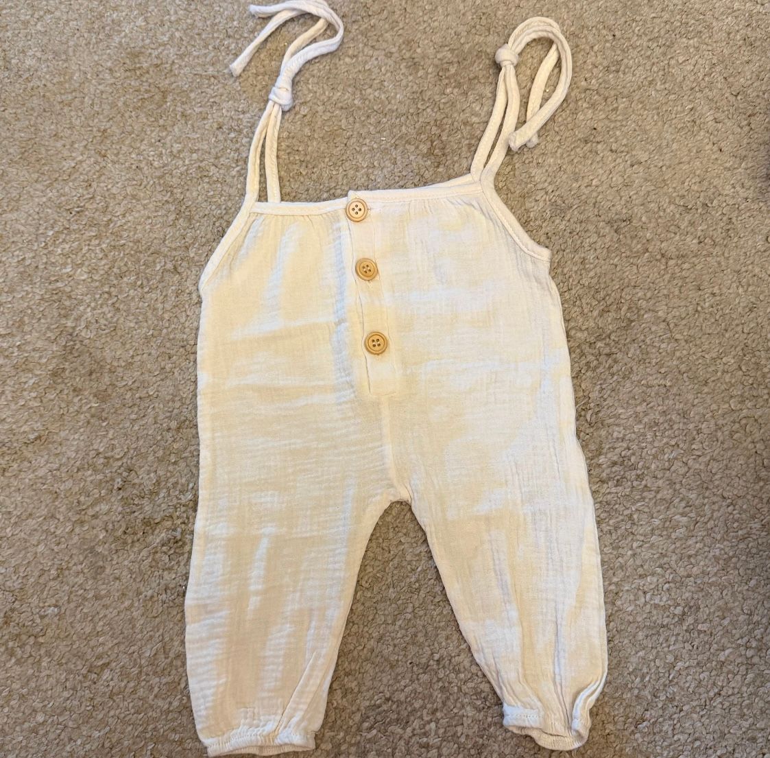 White cotton baby romper with delicate straps and buttons displayed on carpet.