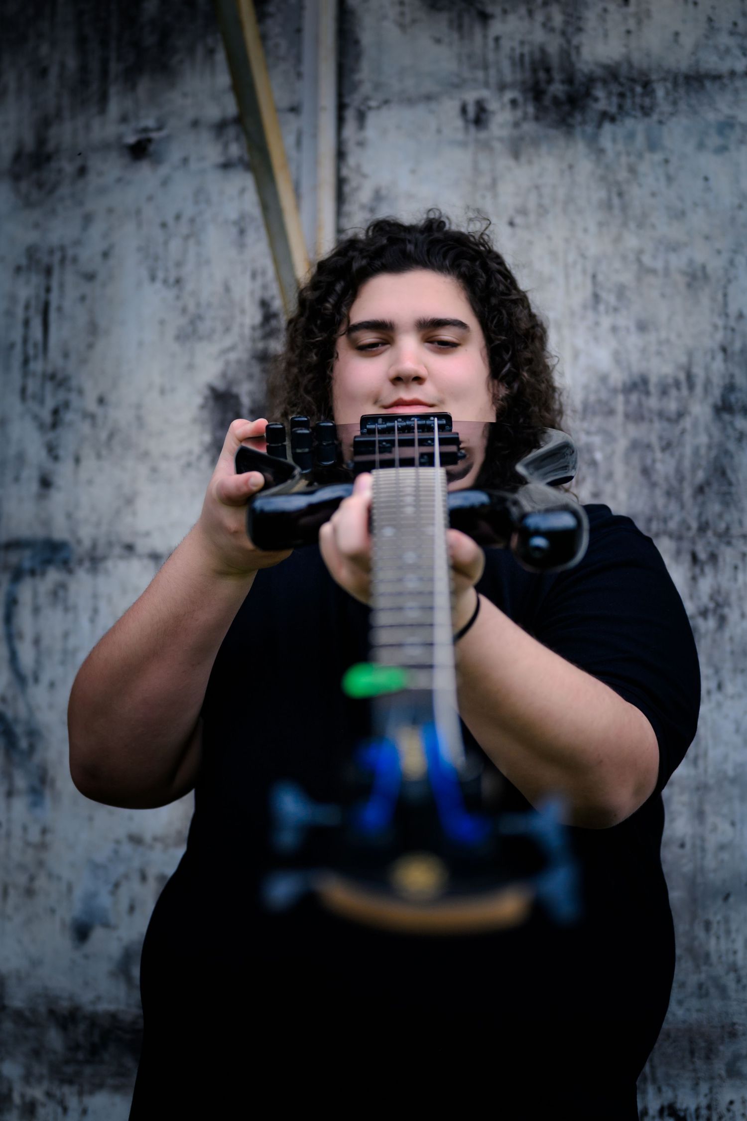 A musician in a black shirt holds a guitar against a textured concrete wall with blurred foreground elements.