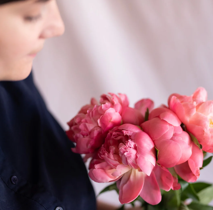 Vibrant coral pink peony flowers arranged in a bouquet against a light background with dark clothing visible.