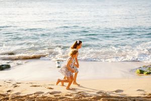 Two young girls in floral dresses running and playing on a sunny sandy beach with ocean waves.