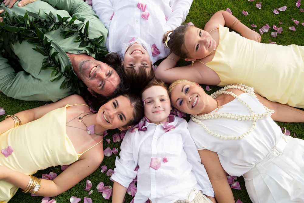 Group lying on grass in a circle formation wearing yellow and white spring outfits with flower petals scattered around them.