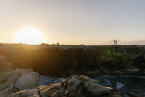 Couple silhouetted against golden sunset on rocky coastal cliffs with ocean waves below.