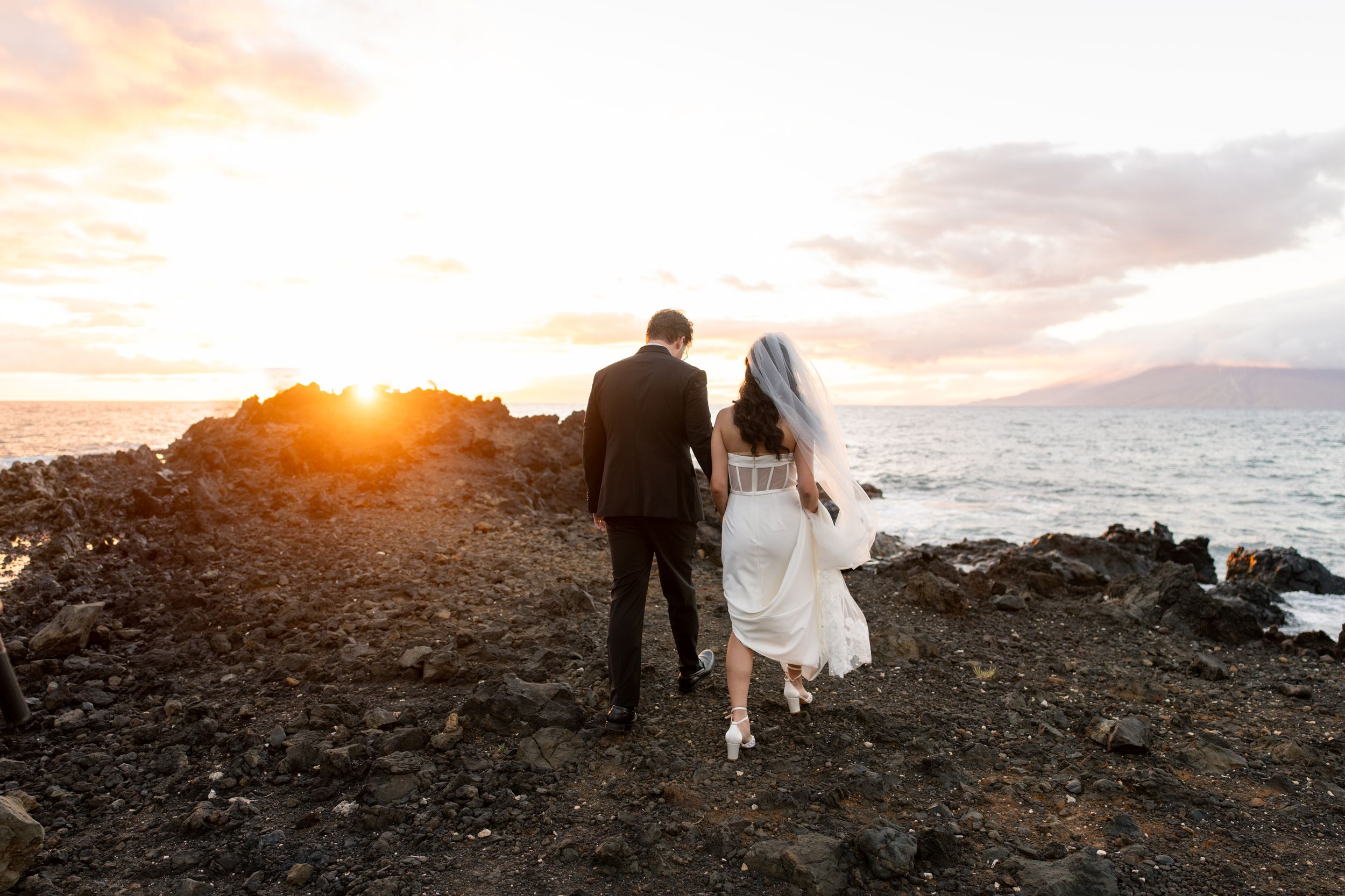 Bride and groom walk hand-in-hand on rocky Hawaiian coastline at golden sunset with ocean and mountains in background.