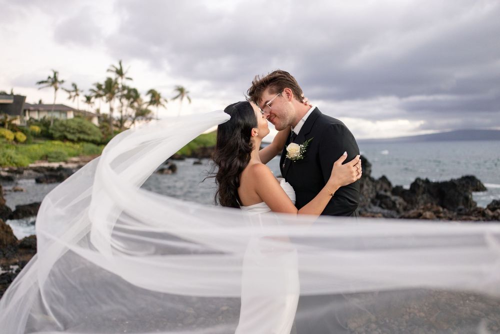 Bride and groom share a romantic kiss on rocky Hawaii shoreline, her veil flowing dramatically in ocean breeze.