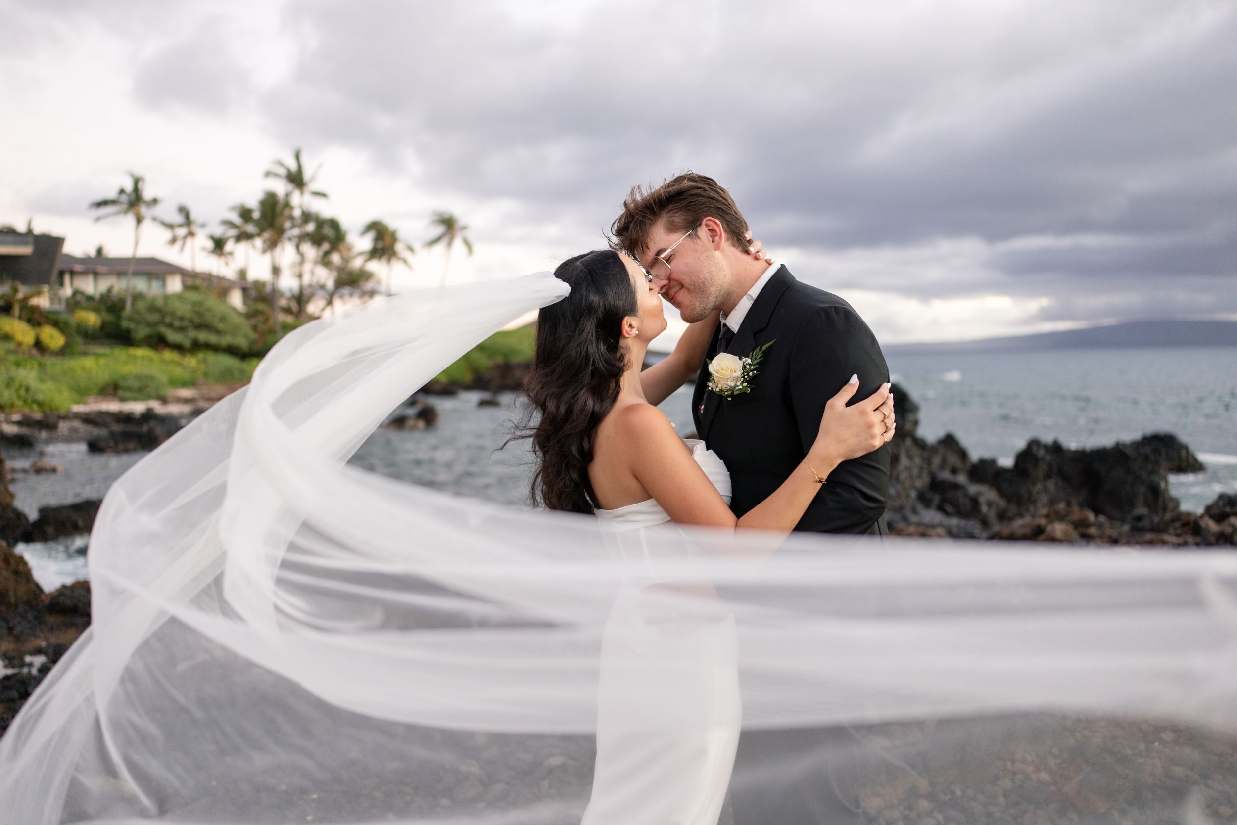 Bride and groom share a romantic kiss on rocky Hawaii shoreline, her veil flowing dramatically in ocean breeze.