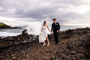 Bride and groom walk hand-in-hand on rocky Hawaiian coastline with dramatic ocean waves crashing behind them.