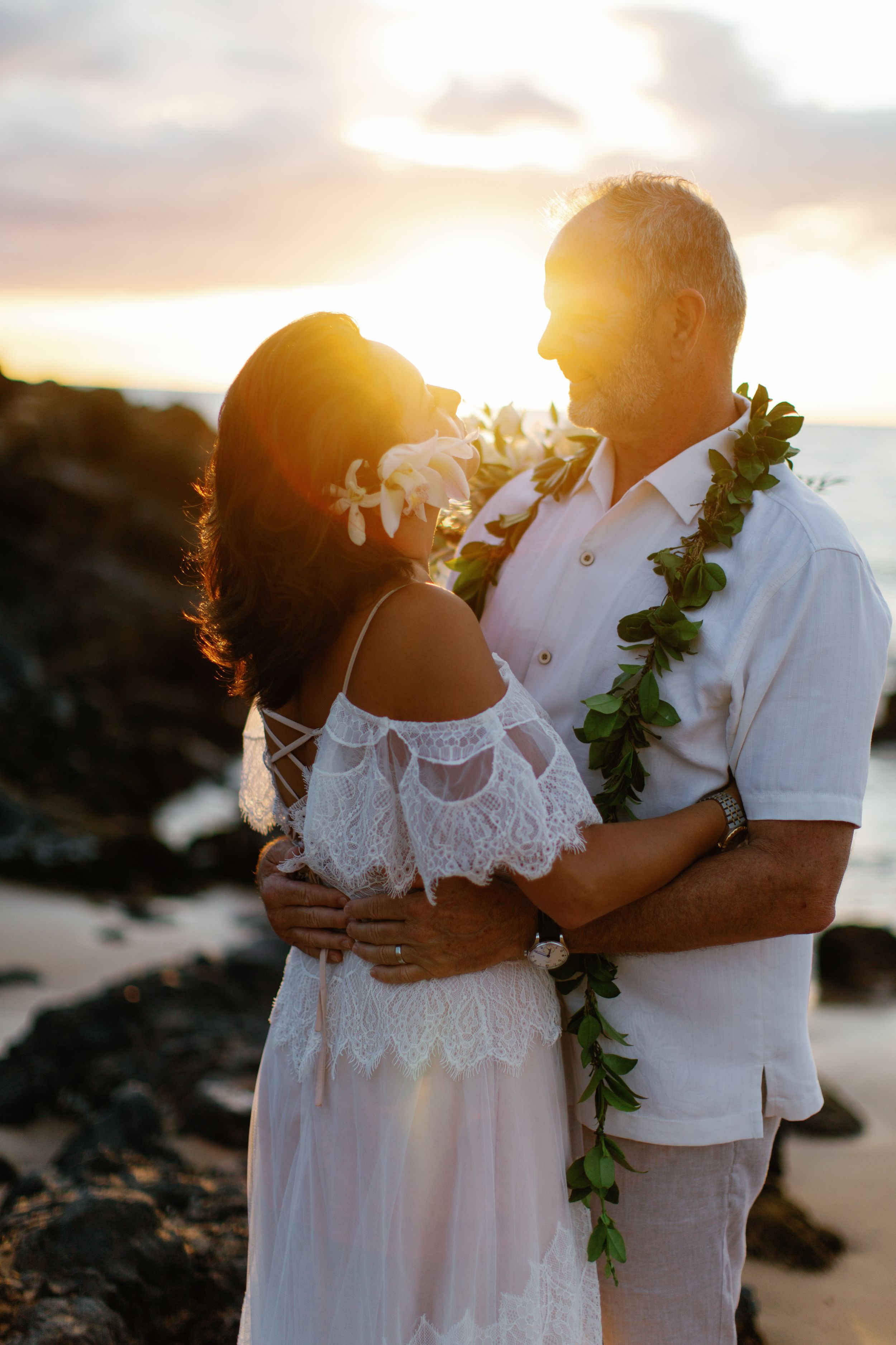 A couple embraces at sunset on a rocky beach in Hawaii, wearing white clothing and traditional lei garland.