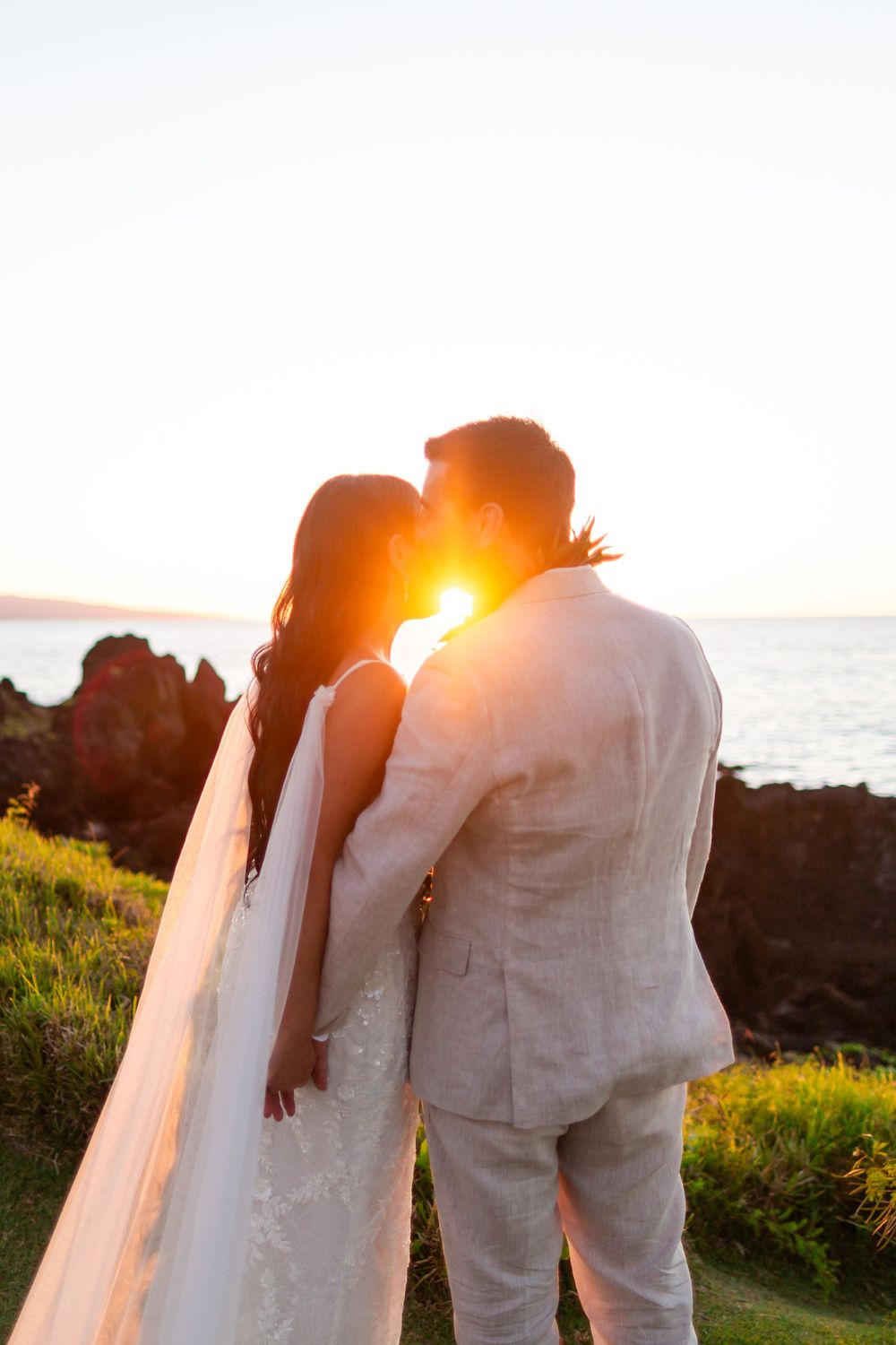 Romantic silhouette of a couple kissing at sunset by the ocean with golden sunlight peeking between them.