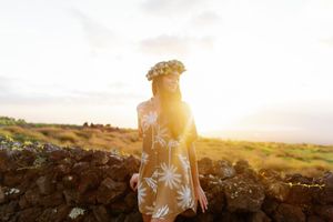 Woman in floral dress and flower crown stands by stone wall at golden sunset in Hawaii.