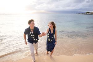 Couple in matching floral outfits laughing and holding hands while walking on a tropical beach at sunset.
