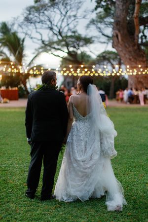 Bride and groom walking hand in hand on green lawn at dusk, with string lights glowing among trees at outdoor wedding reception.
