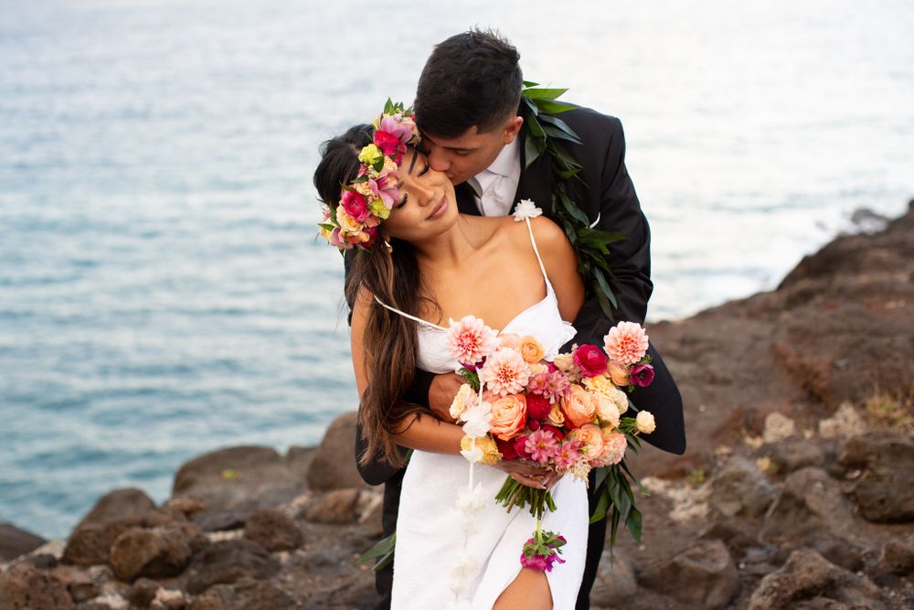 A romantic wedding couple embraces on rocky oceanside cliffs with pink and peach florals and bouquet.