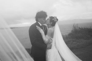 Black and white photo of a bride and groom sharing a romantic kiss outdoors with a flowing veil and scenic landscape.