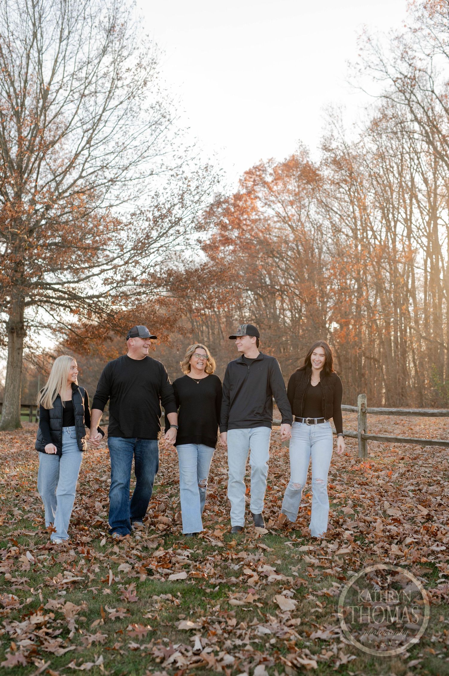 Family walks holding hands along split-rail fence surrounded by fall foliage.