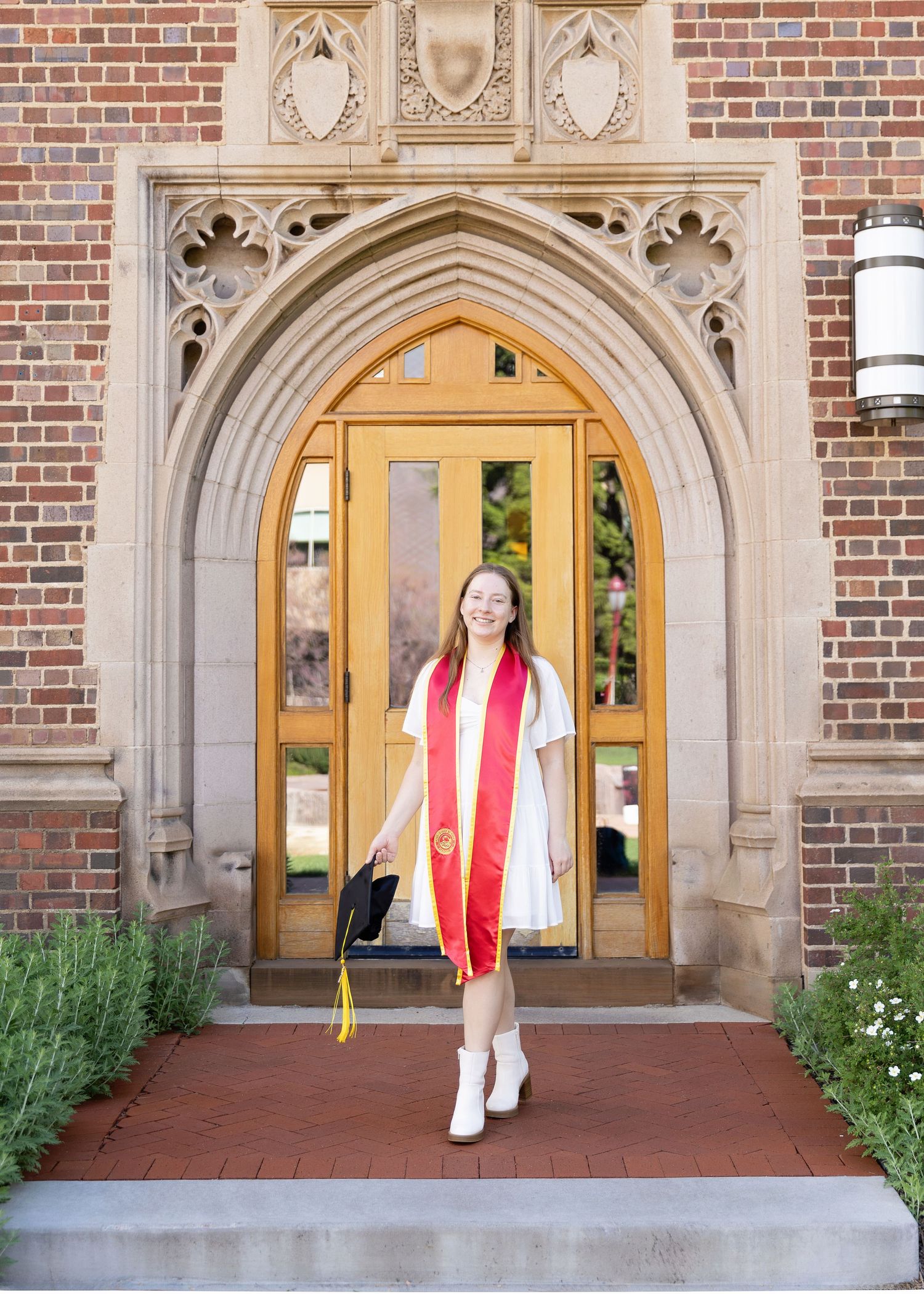 A graduate in white dress and red stole stands in front of an ornate Gothic-style brick archway entrance at DU in Colorado.