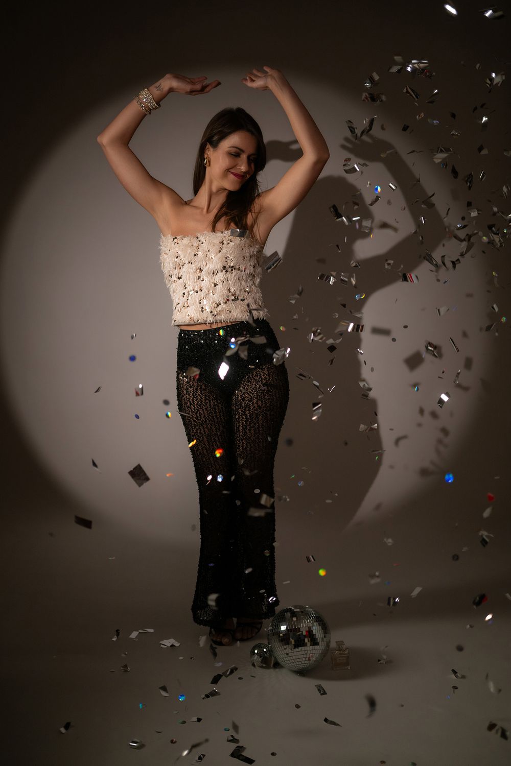 Person dancing in sparkly outfit with confetti falling in dramatic spotlight against dark background.
