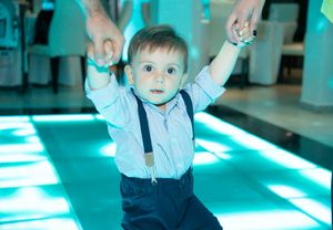 Small child in suspenders and dress shirt dancing under blue party lights.