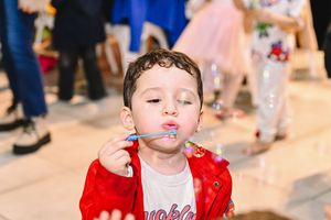 Young boy in red jacket blowing bubbles at a birthday party celebration indoors.