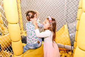 Two young girls playing together in a yellow ball pit play area with safety netting at an indoor playground.