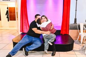 Family of three sharing a warm group hug on a stage with red curtains in a shopping mall.