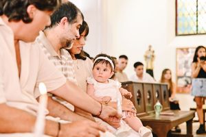 Baby in white dress held by parents during a church baptism ceremony with guests seated in pews.