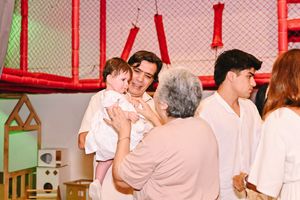 Family gathering indoors with a woman holding a baby, greeted by an elderly lady, while a young man stands nearby.