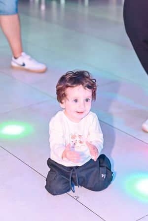 Toddler in white shirt and black pants sits on illuminated dance floor, looking up at camera with colorful lights.