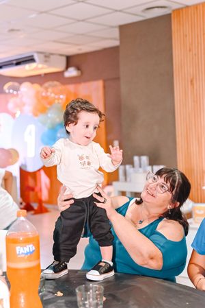 A grandmother joyfully holds a baby standing on a table at a first birthday party with colorful balloons.