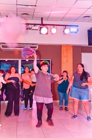 Person with colorful wig holds purple smoke bomb while group dances in vibrant, lit indoor venue.