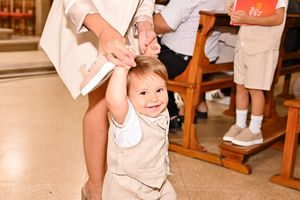 Smiling baby in beige outfit walks with adult help during a church baptism ceremony.