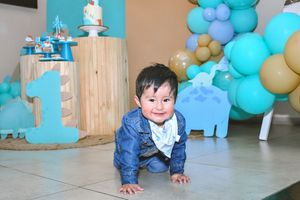 Baby boy in denim jacket crawls on floor at colorful first birthday party with teal balloons and number one decorations.