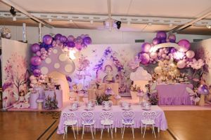 Elegant party setup with purple balloon arch, white furniture and floral decorations.