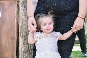 A toddler in a white lace dress holding hands with someone at an outdoor event.