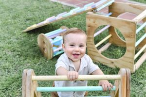 A baby smiling while playing on wooden climbing equipment in a garden setting.