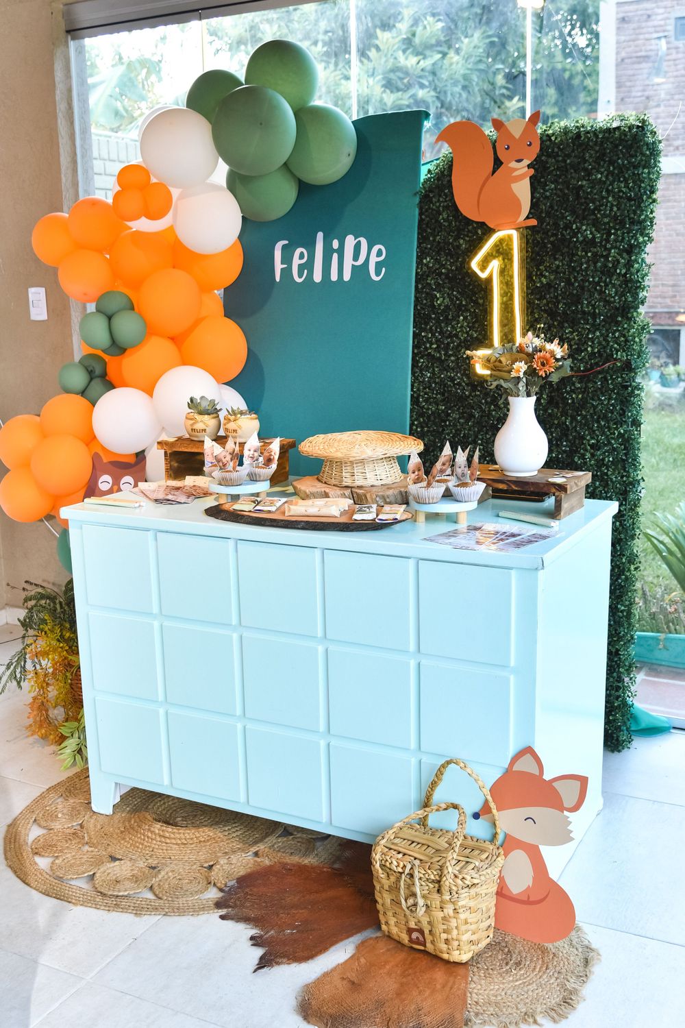 A first birthday party dessert table with orange and green balloon arch and woodland theme decorations.