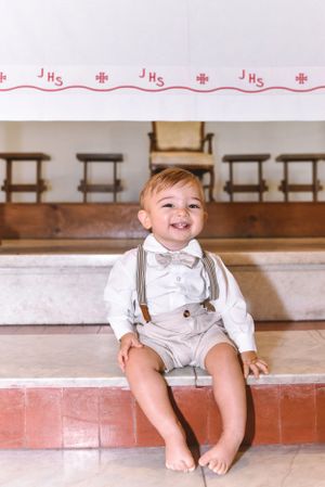 A toddler in formal white attire and suspenders sitting on church steps.