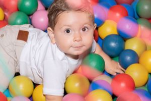 Smiling baby plays in a colorful ball pit filled with pink, blue, red and yellow plastic balls.
