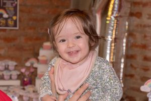 Small child wearing a sparkly silver dress smiles while sitting against a brick wall.