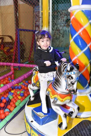 A child rides a colorful carousel horse at an indoor playground with ball pits and inflatable decorations.