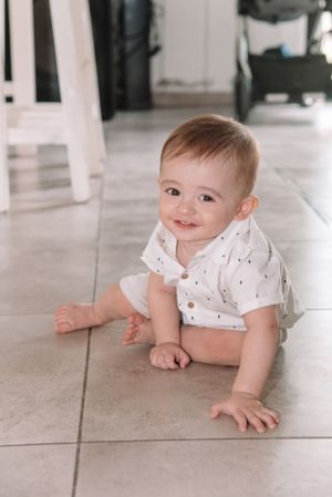 A baby in white clothes crawling on light-colored tile flooring.
