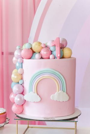 Pink cake decorated with pastel rainbow designs, clouds, and colorful ball ornaments on a gold stand.