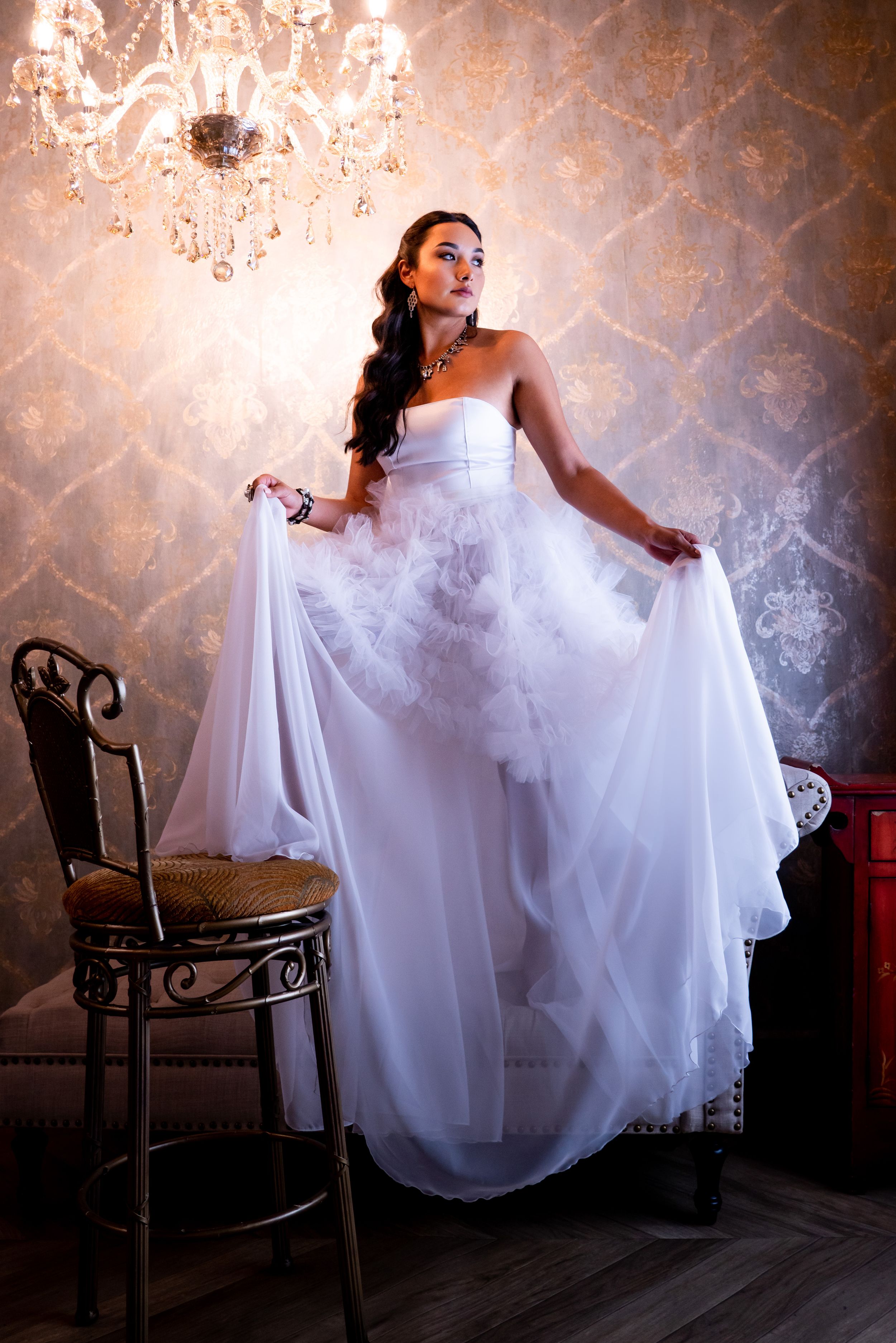 Elegant bride in white gown poses by vintage chandelier and patterned wallpaper, during a bridal portrait session.